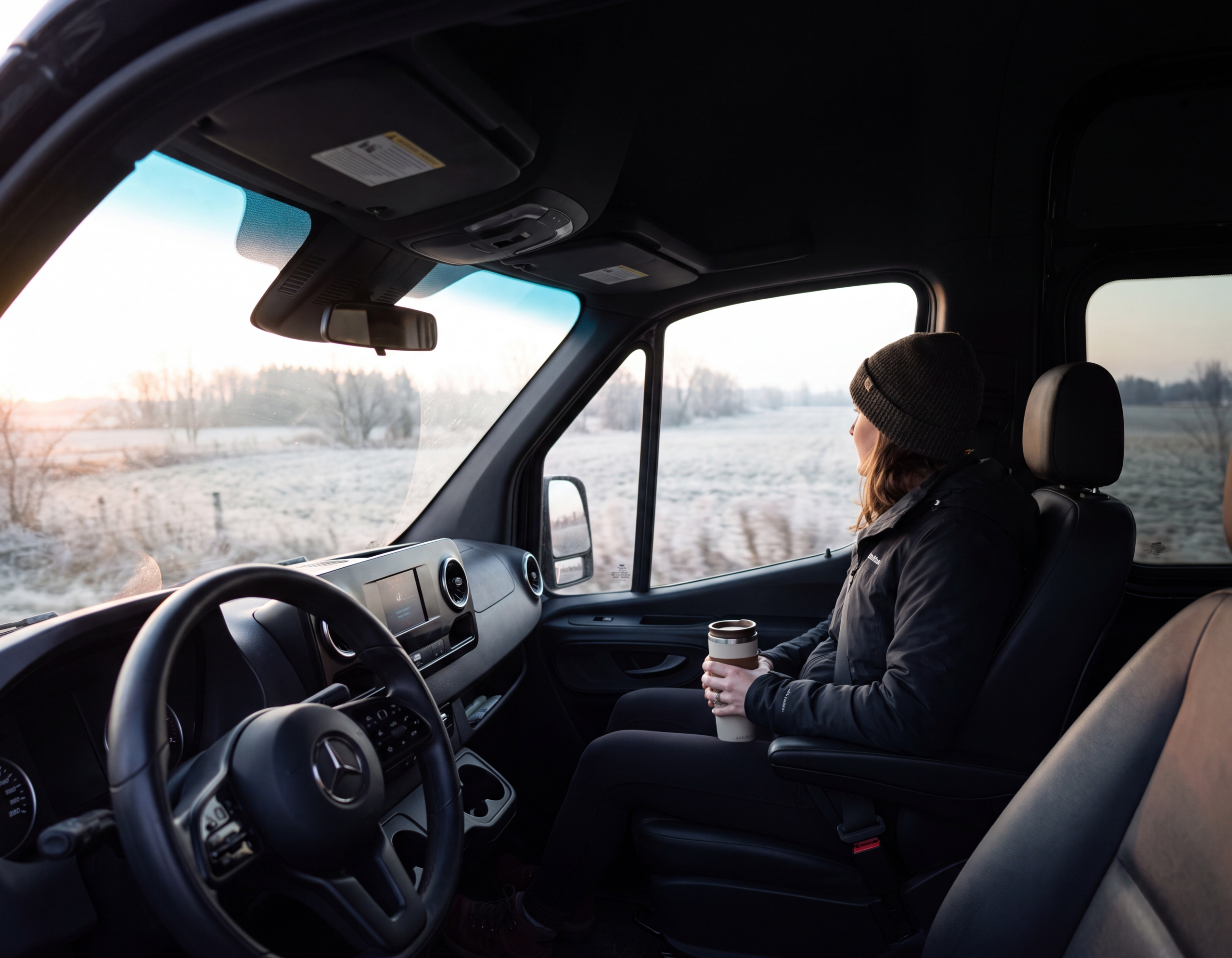Interior of a black Mercedes-Benz Sprinter cabin, one passenger seated with a coffee cup looking out the window during an early-morning highway run near Belleville Ontario