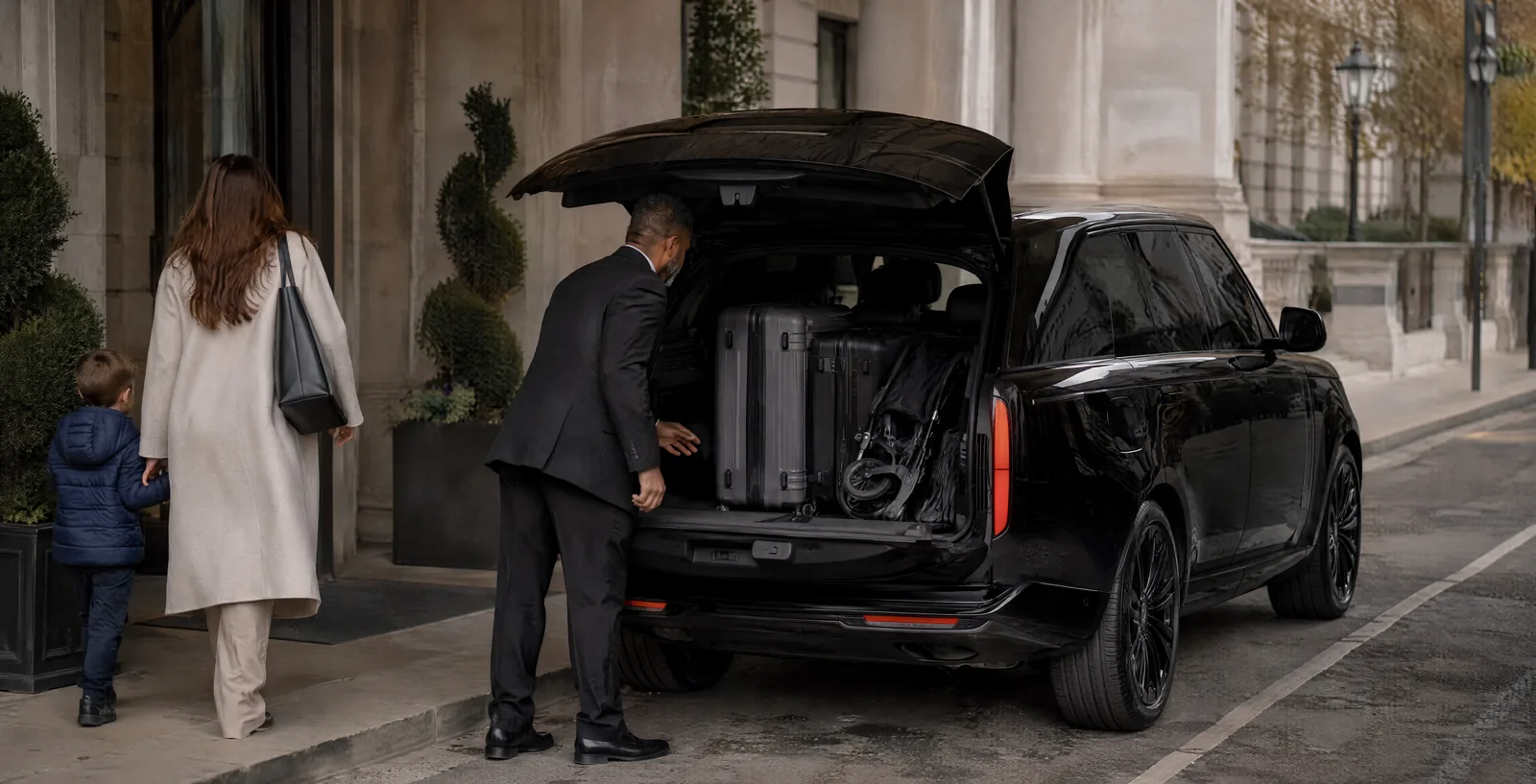 A chauffeur loading a family's luggage and a stroller into a black SUV trunk at a hotel kerb