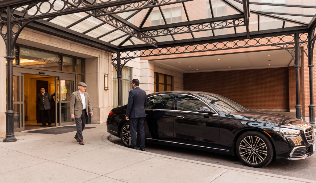 RM Locations 39 Chauffeuropolis chauffeur holding the rear door of a black car at the kerb for an arriving passenger, hero residence portico morning